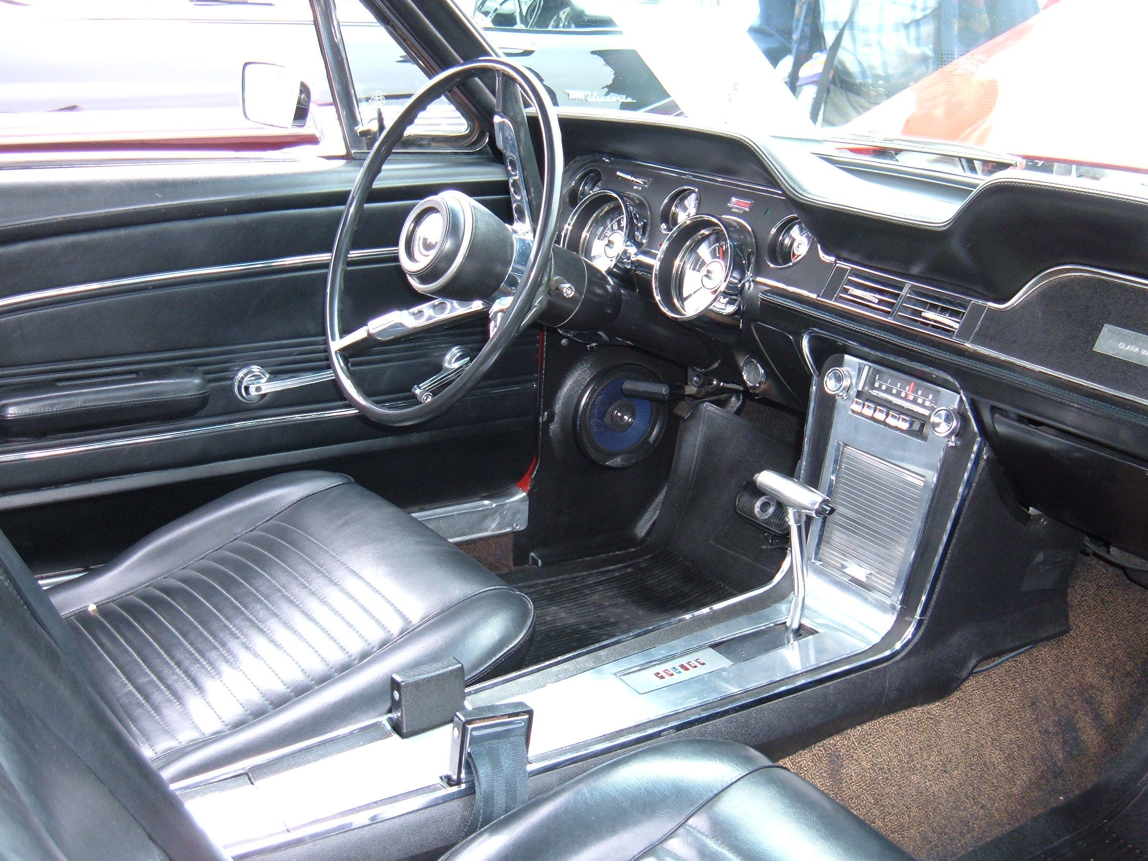 Real 1967 Mustang coupe interior showing the pleated black seat cushion at lower left, the left door panel and armrest, and the dashboard and console.